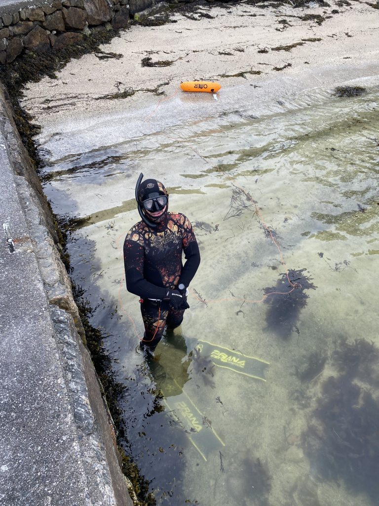 En mann kledd i vptdrakt, med dykkermaske og snorkel på, står klar til å gå ut i vannet fra en sandstrand
